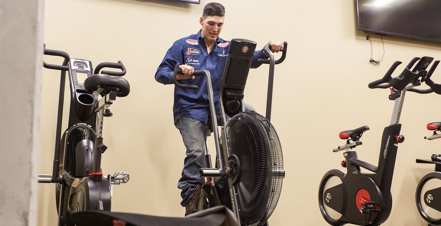 Jess Pope lleva una camisa azul abotonada y pantalones vaqueros azules, y monta una bicicleta fija para calentar antes de competir en un rodeo.